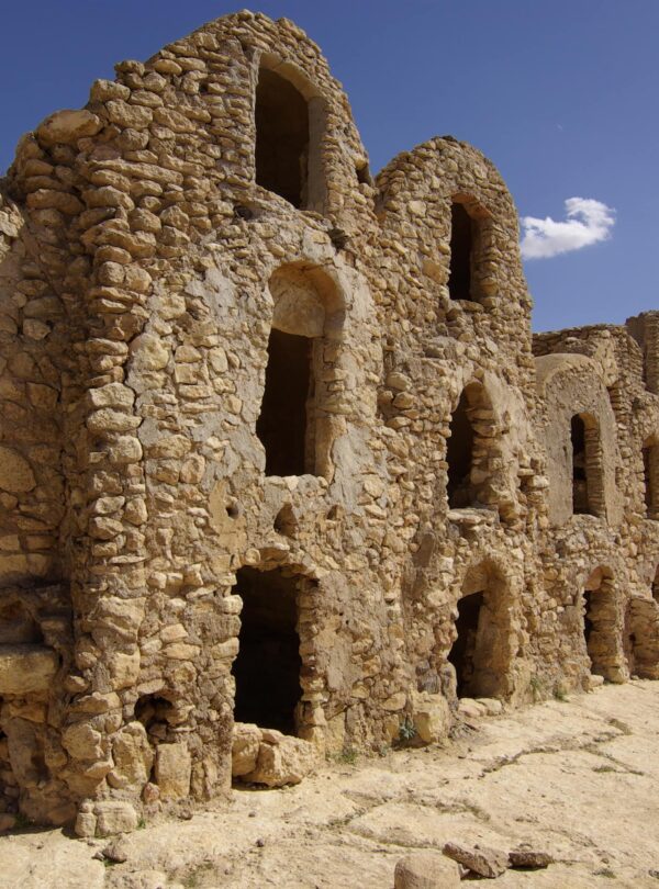 Vue aérienne de Ksar Jouamaâ, perchée sur un piton escarpé dans le gouvernorat de Médenine, Tunisie. Les structures rectangulaires irrégulières du ksar, divisées en sections anciennes et modernes, révèlent une mosaïque de ghorfas restaurées et en ruine, témoignant de l'architecture berbère traditionnelle.